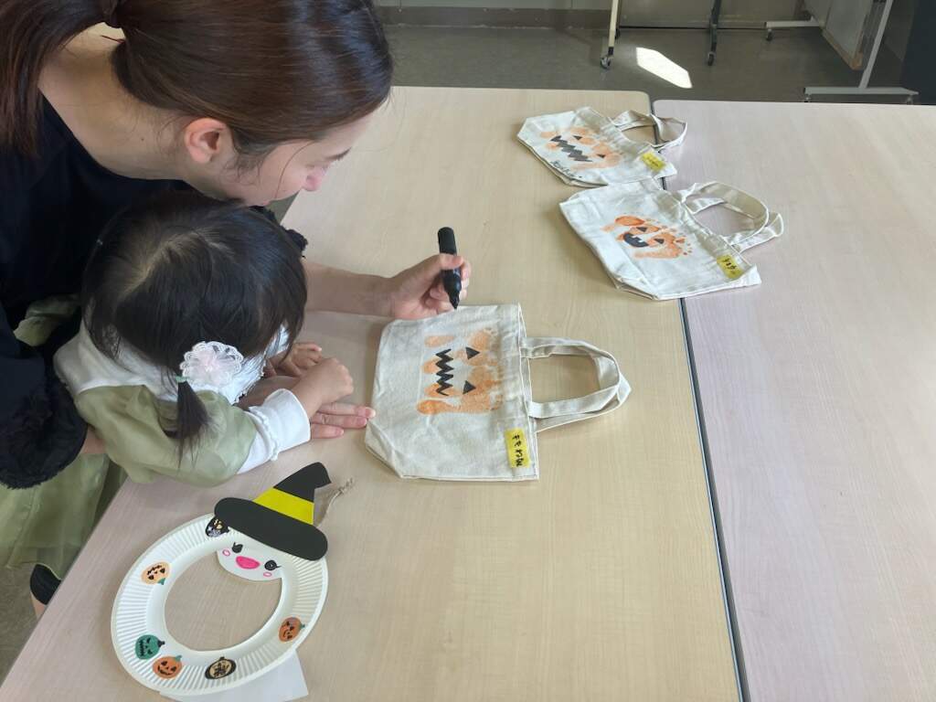 お母さんと女の子が小さいトートバックにオレンジ色の絵の具でカボチャ柄になるように足型スタンプを押して制作している様子