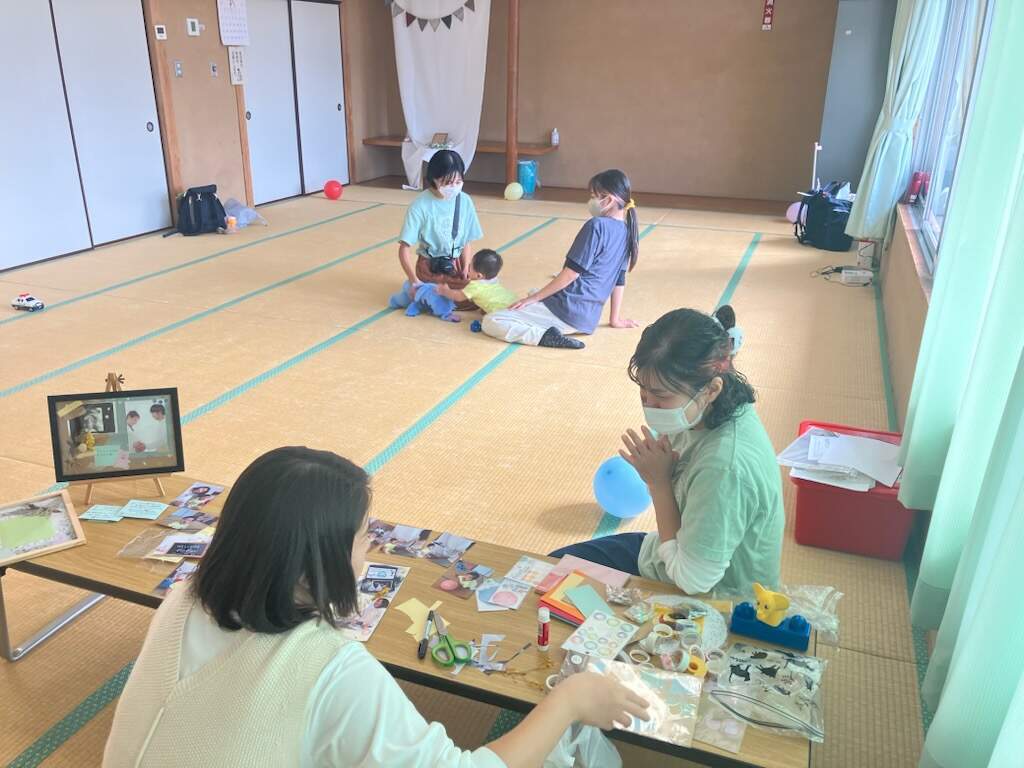 参加したお母さんがスタッフと談笑している後ろでこどもとスタッフが遊んでいる様子