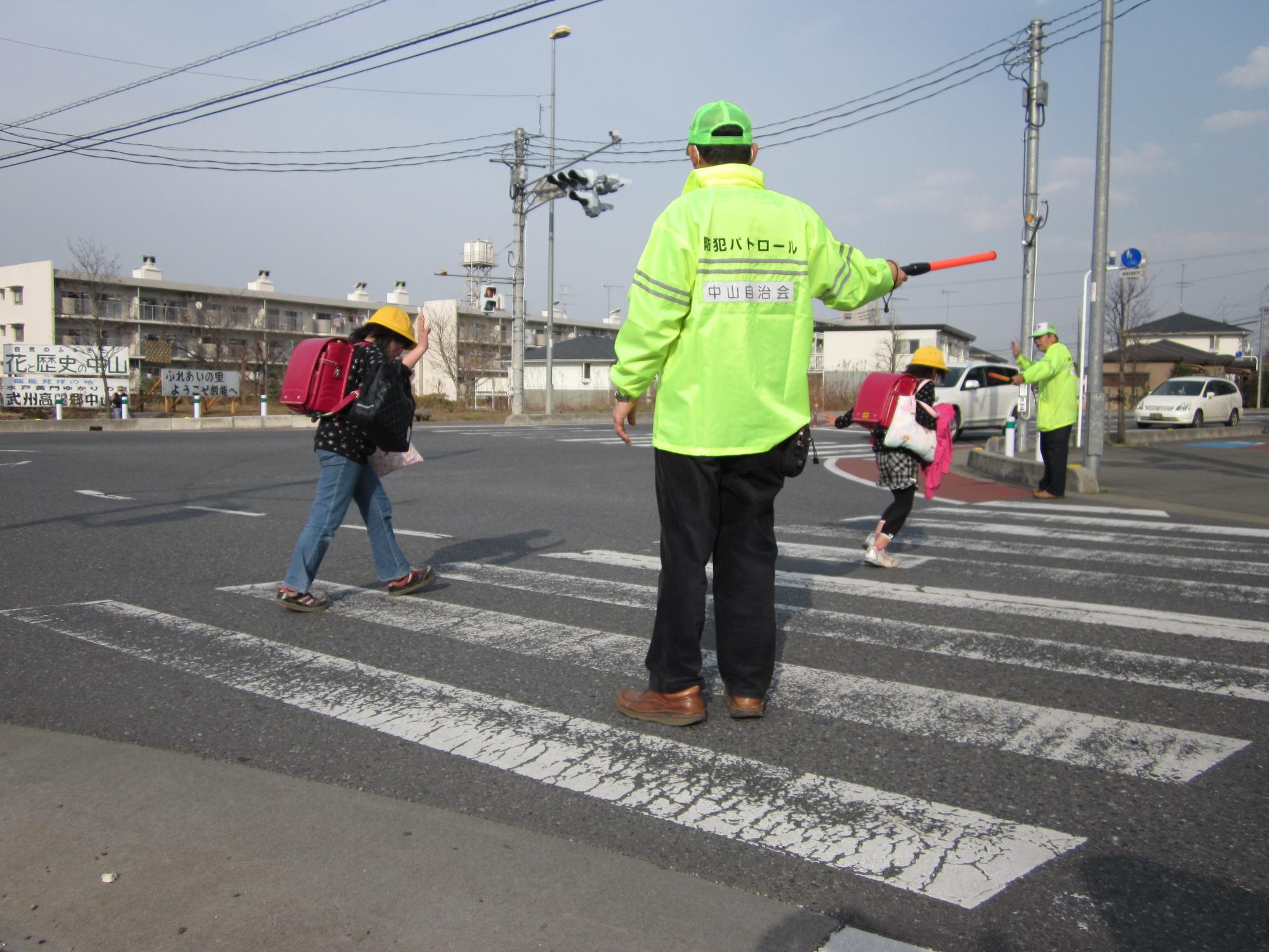 横断歩道で下校する児童らを見守る中山自治会自主防犯グループ隊員らの写真
