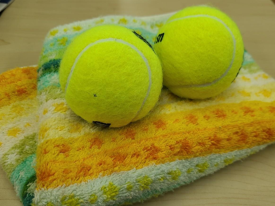 タオルとテニスボールの写真。オレンジ色、白、緑のストライプ柄の畳んだタオルの上に、硬式の黄色いテニスボールが2個乗っています。