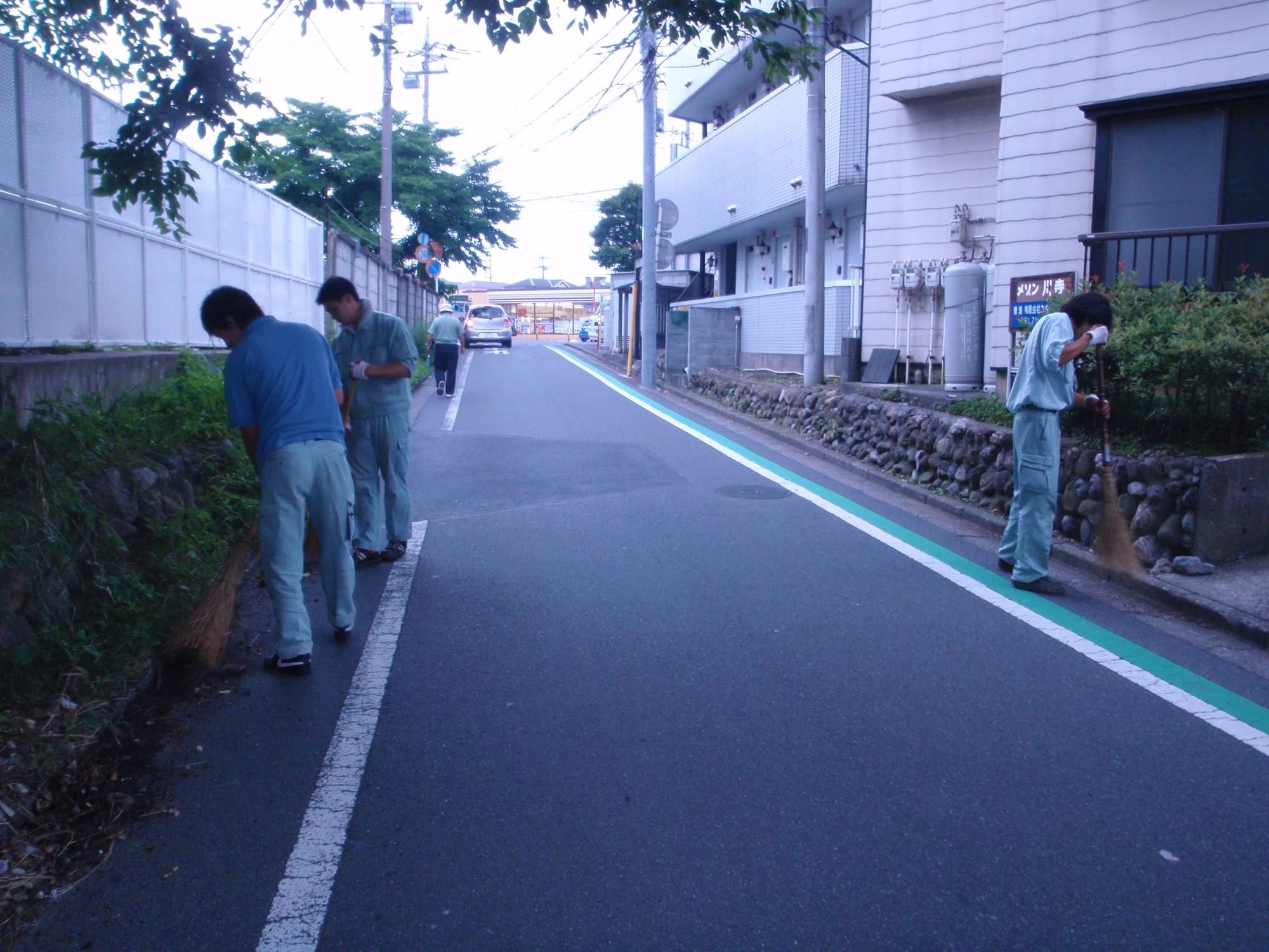 有限会社飯能清掃センターの方々が、ほうきを使い手分けして道路脇の清掃作業を行っている様子の写真