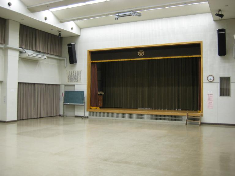 天井が高く、舞台があり黒板が設置されている集会室の写真