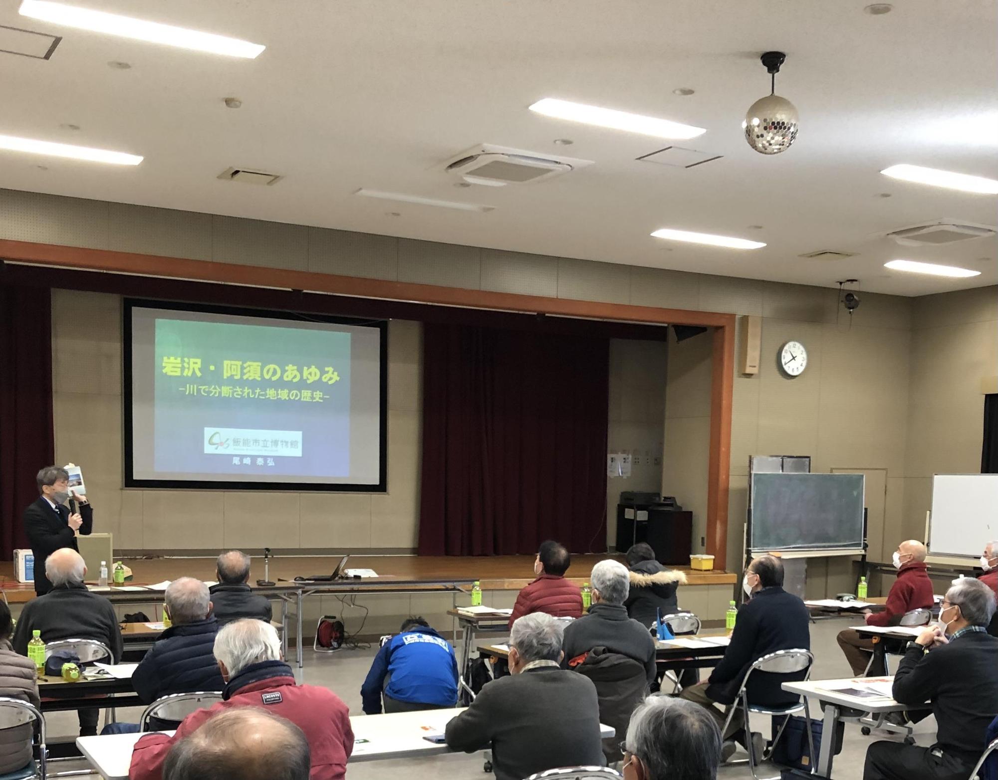 講座参加者に岩沢と阿須の歴史についてスクリーンで説明をしている尾崎館長の写真