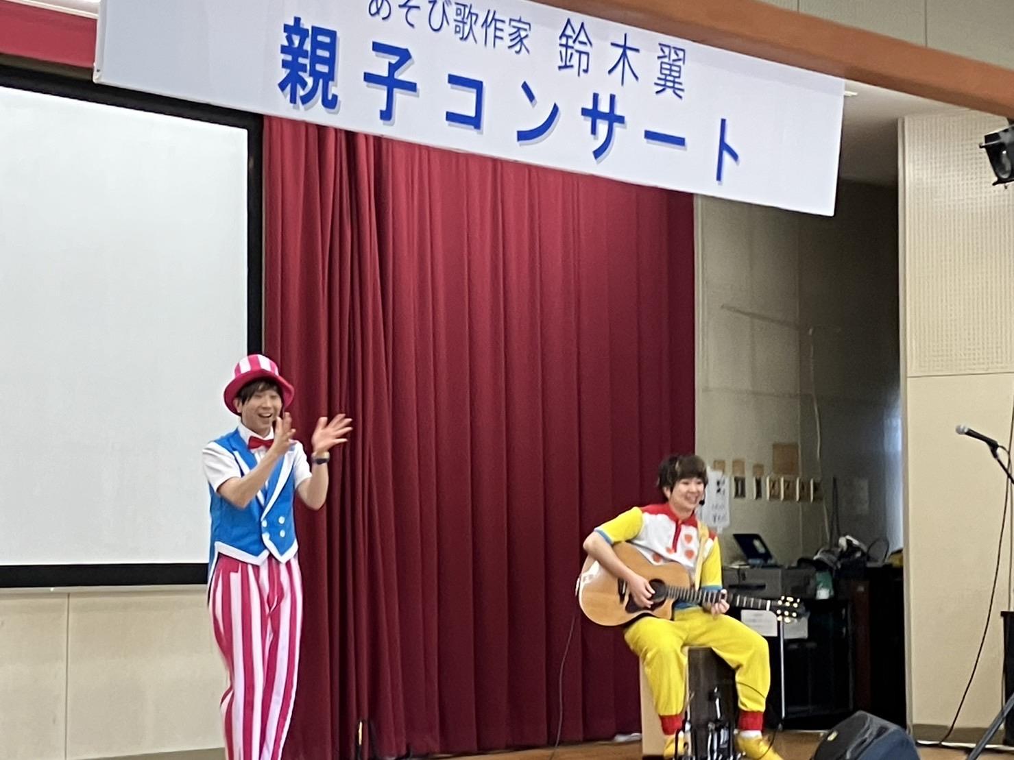 舞台上で遊び歌作家鈴木翼さんがピンクと白のストライブプの防止とズボンに水色のベストに赤い蝶ネクタイをして手拍子しながら歌いその隣で黄色い服を着た男性が座ってギターを弾きながら伴奏する様子。