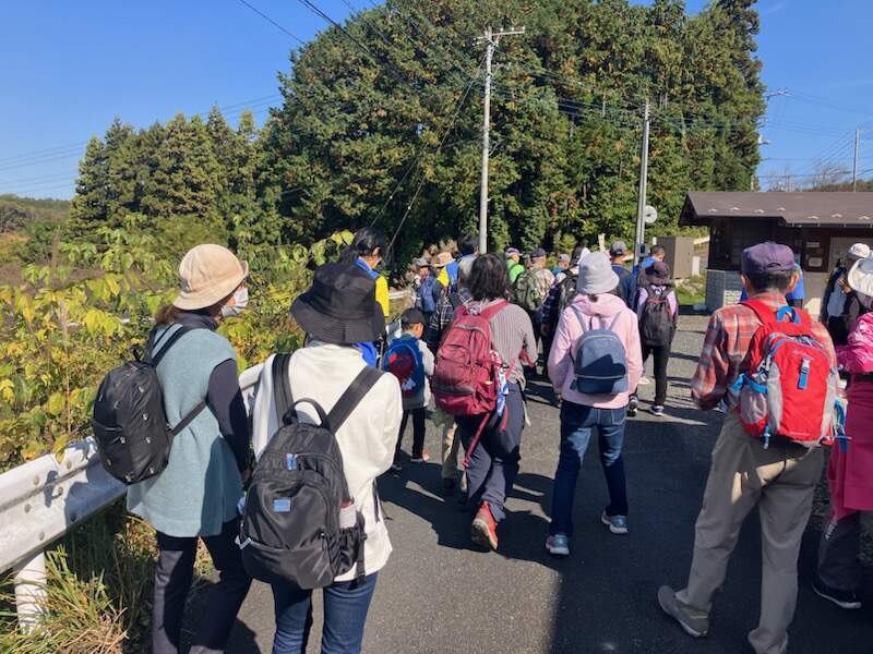 秋晴れの青空の下、リュックを背負った参加者が談笑しながら歩いている様子