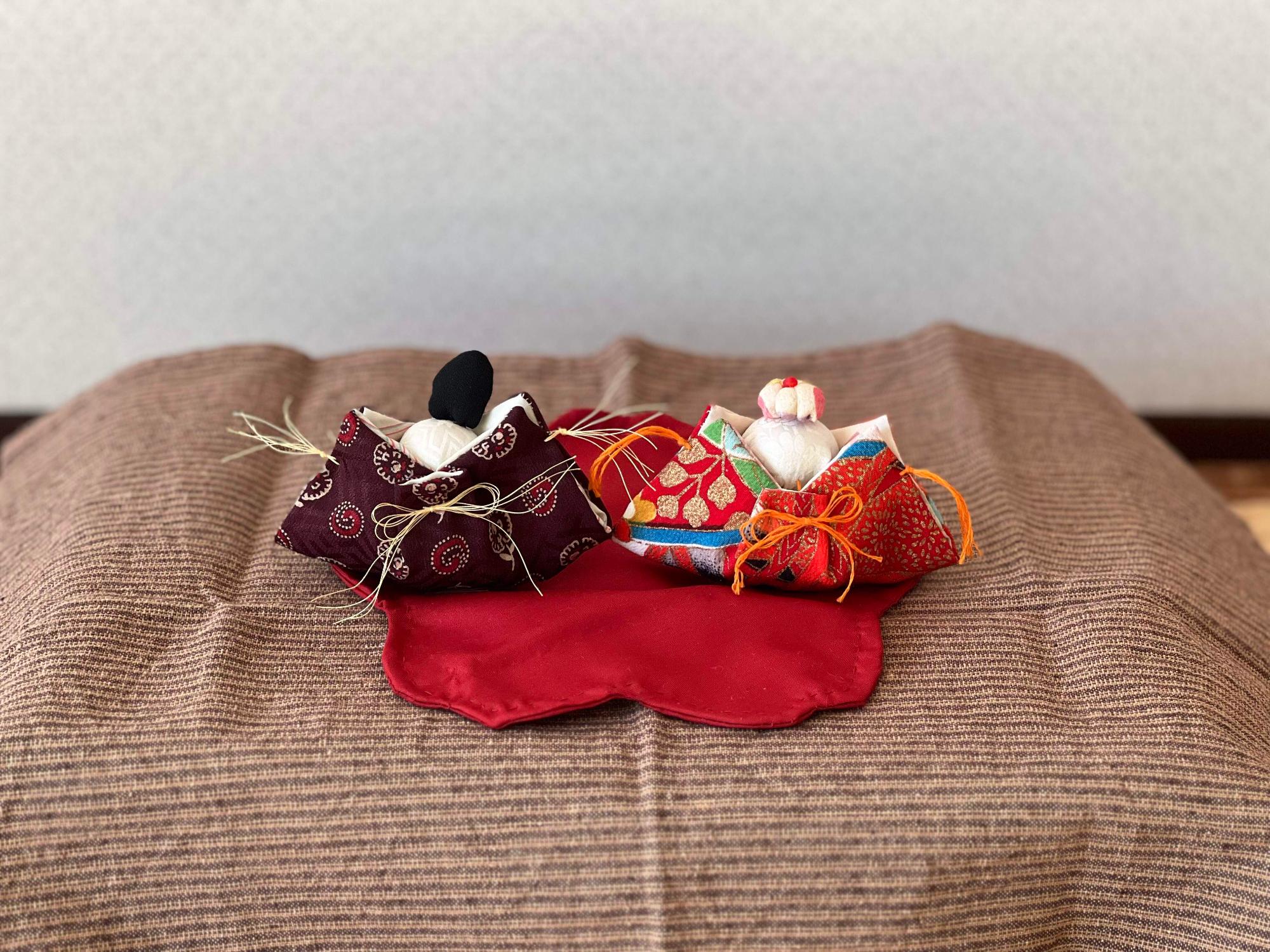 えんじ色の花びらのマットの上に6センチくらいの四角い着物布を半分に折り畳んだ上に白い頭がついたお内裏様とおひな様。お内裏様の頭には黒い冠、おひな様には着物布で作った冠がのっている。