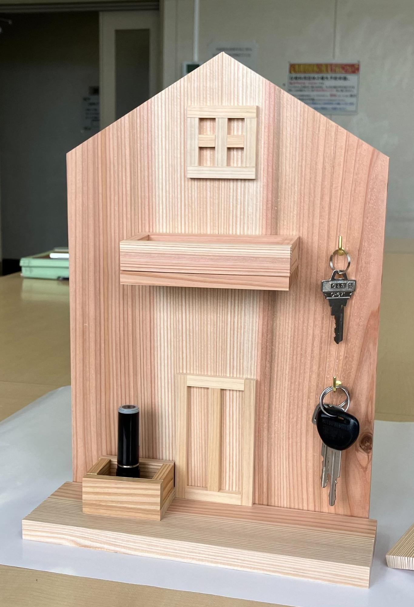 木工作広場で作ったキーラック。西川材の板を家の形に切ってあり、そこに薄く小さい木片で窓やドアを貼り付けてあったり、箱のような入れ物もついている。また、鍵をひっかけるL字フックもついている。